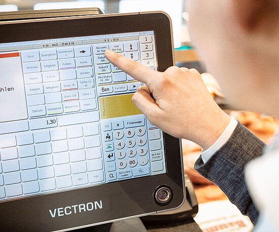 Eine Person bedient eine Vectron-Kasse und tippt auf den Touchscreen, auf dem verschiedene Verkaufsoptionen und Preise angezeigt werden.