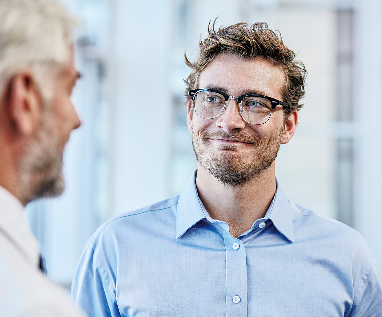 Ein junger Mann mit Brille und Bart, der ein hellblaues Hemd trägt, lächelt und schaut einen älteren Mann mit grauen Haaren an. Der Hintergrund ist unscharf und zeigt ein Büro oder eine Arbeitsumgebung.