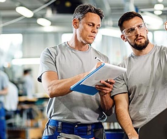 Zwei Männer in einer Fabrik nehmen sich Notizen und schauen sich ein Teilchen an