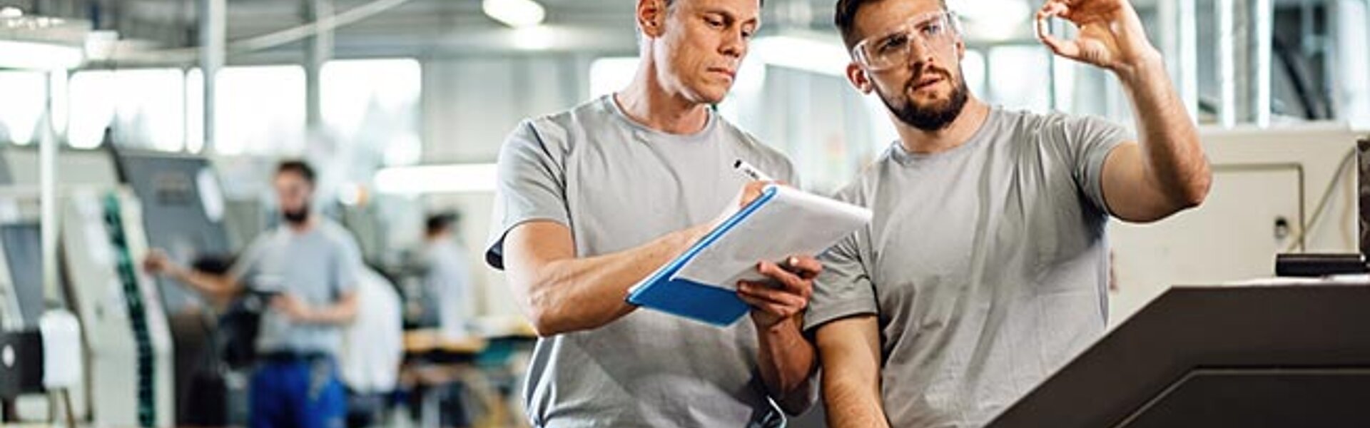 Zwei Männer in einer Fabrik nehmen sich Notizen und schauen sich ein Teilchen an