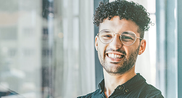 Junger Mann mit Brille steht vor Fensterfront und lacht