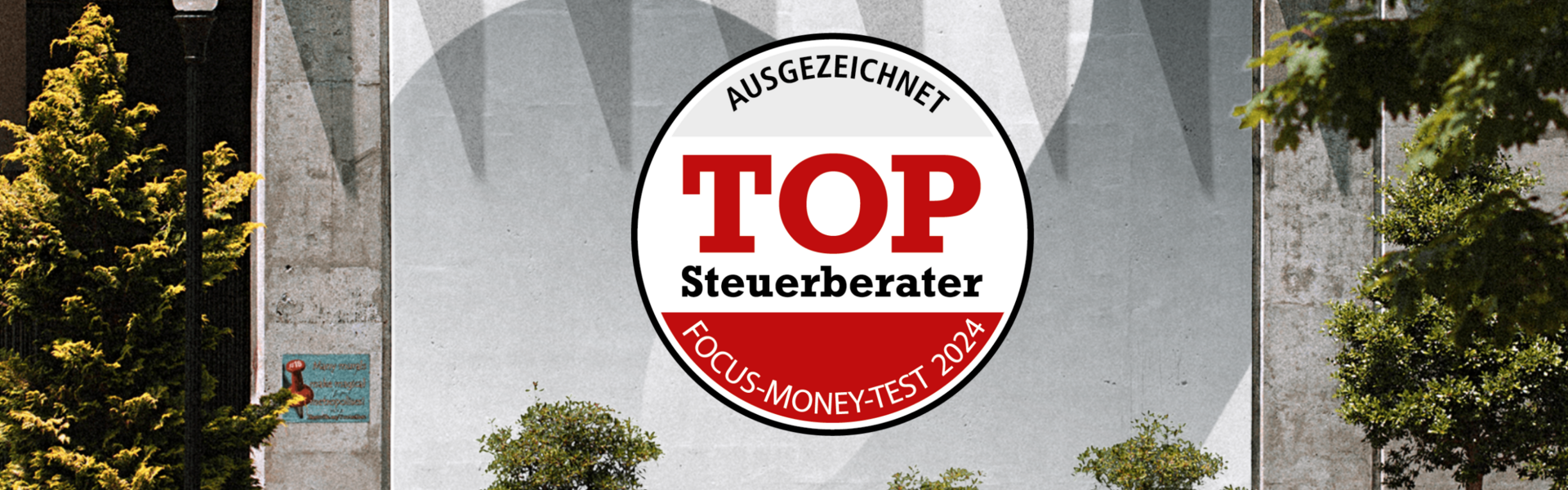 Ein großes Plakat an einer Betonwand mit der Aufschrift "Ausgezeichnet TOP Steuerberater Focus-Money-Test 2024", umgeben von Pflanzen und einem Gehweg.