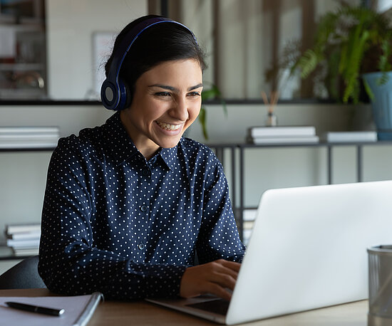 Frau sitzt am Schreibtisch mit Kopfhörern und schaut enthusiastisch auf Laptop