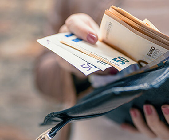 Eine Person hält eine geöffnete schwarze Geldbörse, aus der mehrere Euro-Banknoten, darunter 50-Euro-Scheine, herausragen. Der Hintergrund ist unscharf.