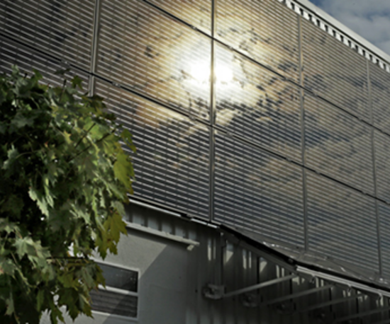 Eine Reihe von Solarpaneelen an der Fassade eines Gebäudes, die das Sonnenlicht reflektieren, mit einem Baum links im Bild.