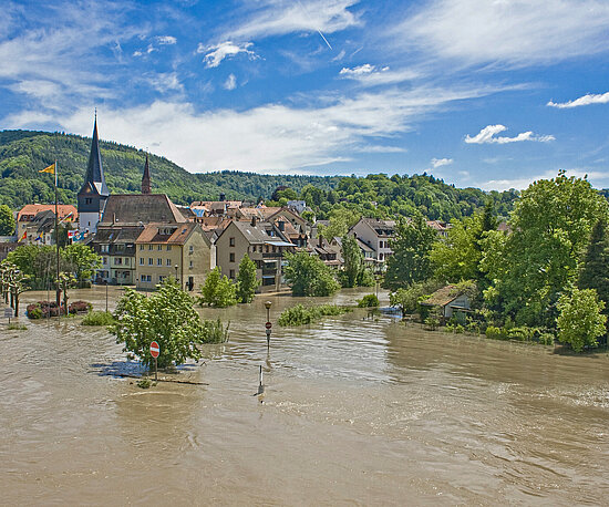 Eine überschwemmte Stadt mit einer Kirche und mehreren Häusern. Das Wasser bedeckt Straßen und Bäume, während die Hügel im Hintergrund und der blaue Himmel eine idyllische Kulisse bilden.